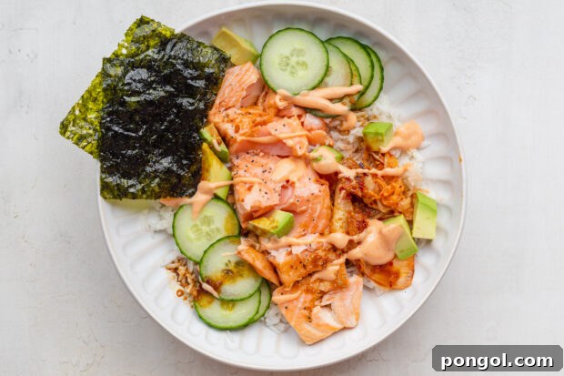 Overhead view of a salmon rice bowl with cucumber, avocado, kimchi, and nori, topped with sriracha mayo and soy sauce.