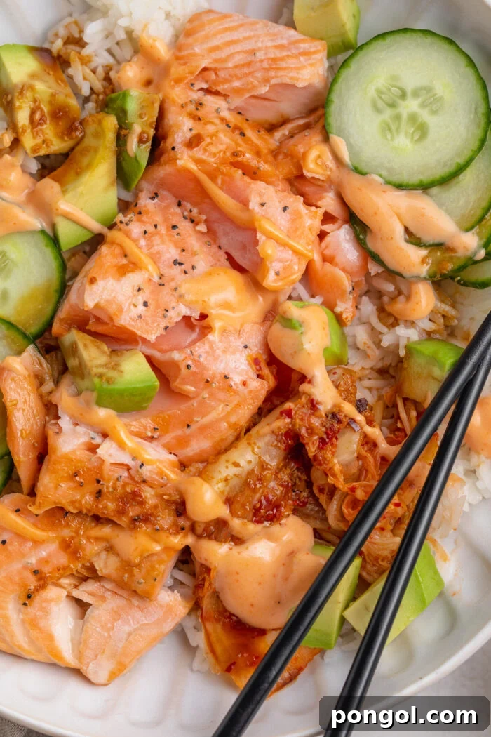 Close-up view of oven-baked salmon fillets with white rice, soy sauce, creamy sriracha mayo, cucumber, avocado, and chopsticks.
