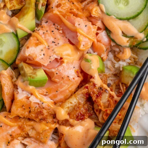 Close-up view of oven-baked salmon fillets with white rice, soy sauce, creamy sriracha mayo, cucumber, avocado, and chopsticks.