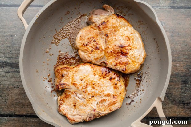 Overhead view of two pan-seared pork chops in a large skillet on a wooden tabletop.