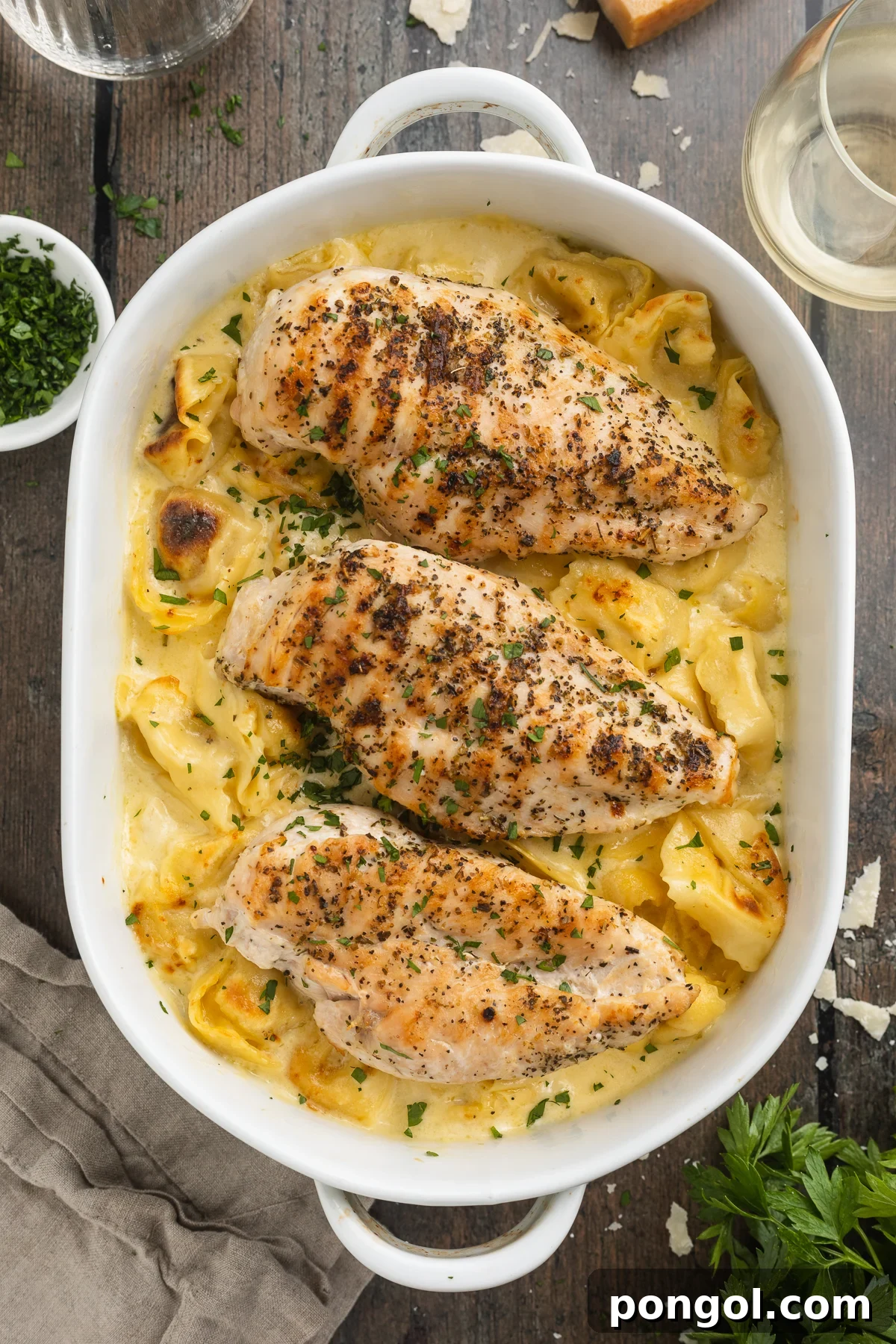 Overhead view of a large oval white baking dish holding copycat asiago tortelloni alfredo topped with 3 grilled chicken breasts.