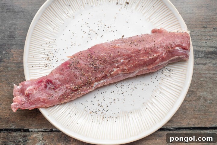 Overhead view of uncooked pork tenderloin, seasoned with salt and black pepper, resting on a white plate on a wooden table.