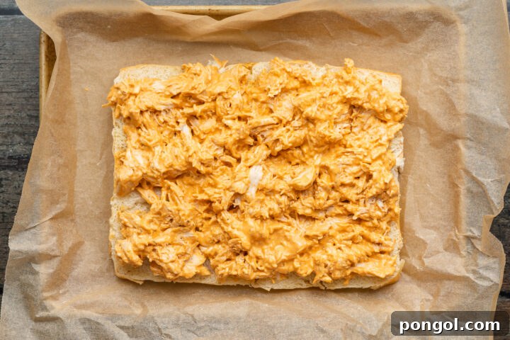 Spicy Buffalo Chicken Minis 7 Overhead view of buffalo chicken spread out on top of a slab of halved sweet dinner rolls in a baking dish lined with parchment paper.