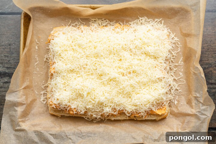 Spicy Buffalo Chicken Minis 8 Overhead view of buffalo chicken topped with mozzarella, spread out on top of a slab of halved sweet dinner rolls in a baking dish lined with parchment paper.