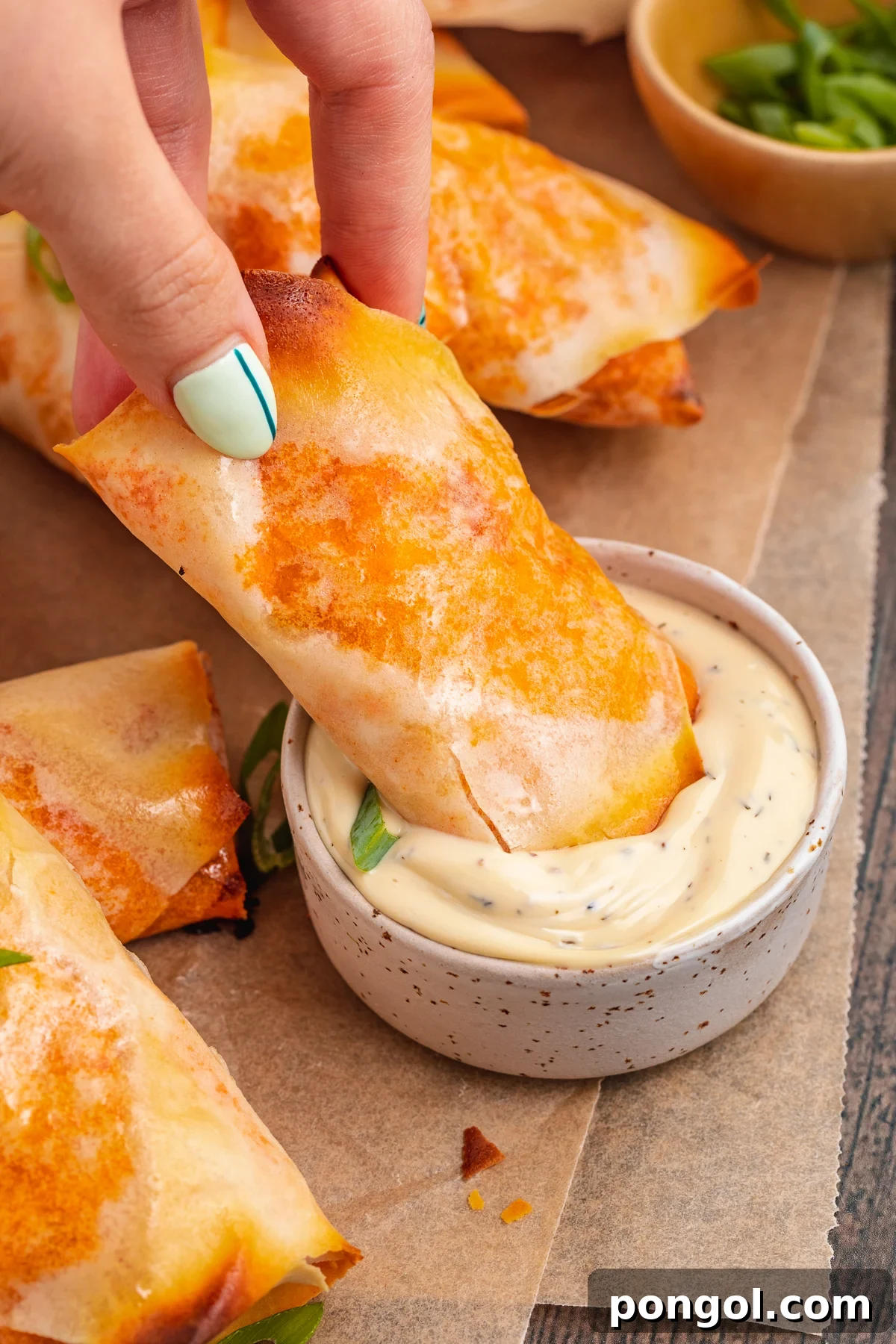 A white woman's hand with painted nails dipping a buffalo chicken egg roll into a small ramekin of ranch dressing on a baking sheet lined with parchment paper.