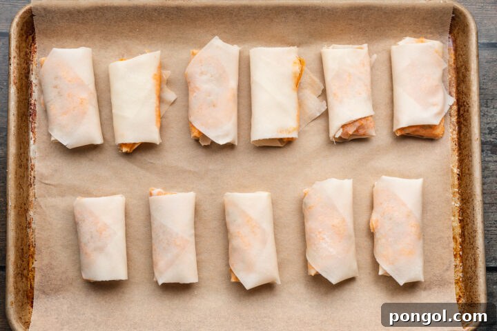 Overhead view of rolled and sealed buffalo chicken egg roll wrappers on a baking sheet lined with parchment paper.