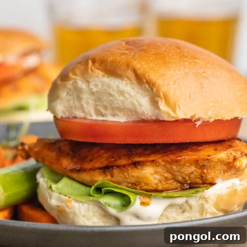 Zesty Buffalo Chicken Stack 4 Side view of a buffalo chicken sandwich with an orange-colored grilled chicken breast, a thick slice of tomato, green lettuce, and a fluffy bun on a blue-grey plate.