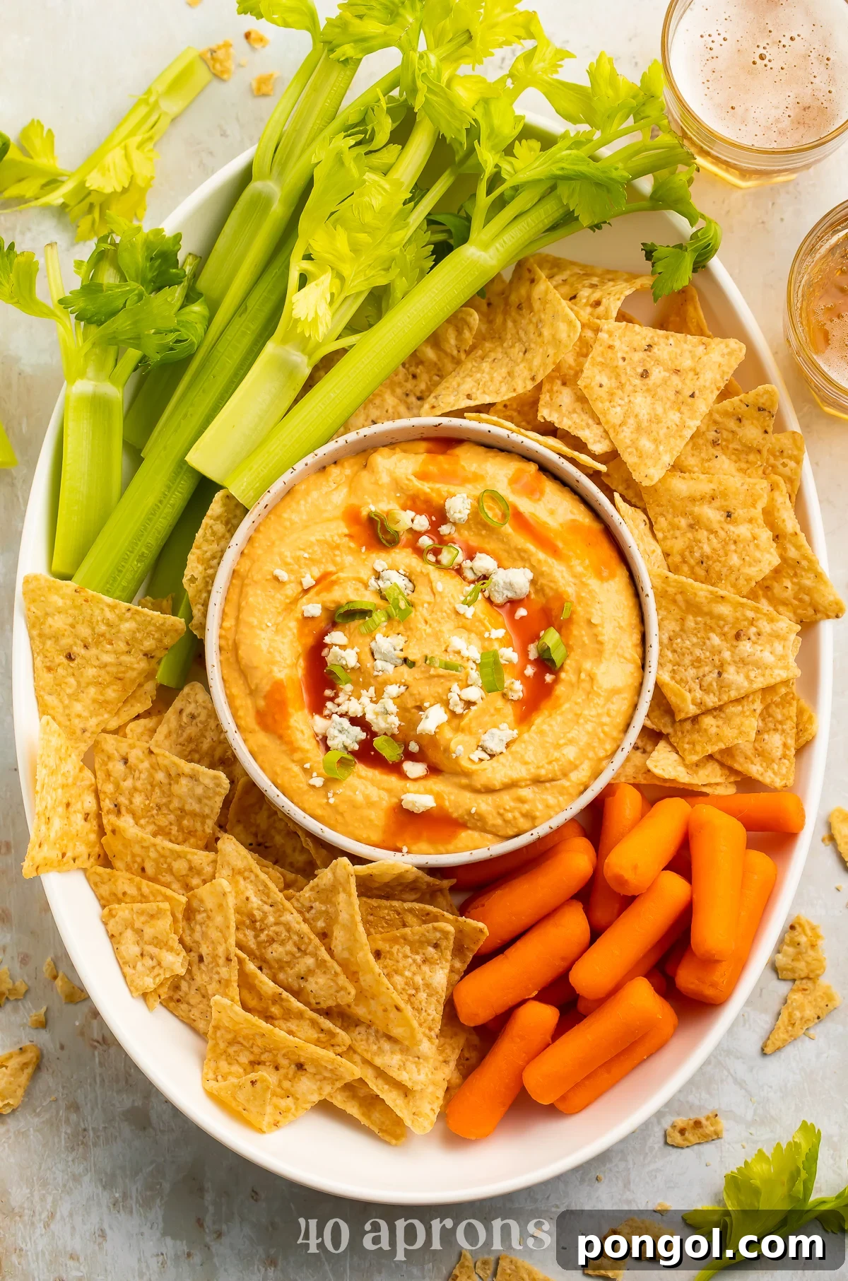 Overhead view of a bowl of buffalo hummus topped with blue cheese crumbles in the center of a platter surrounded by celery, carrots, and tortilla chips.
