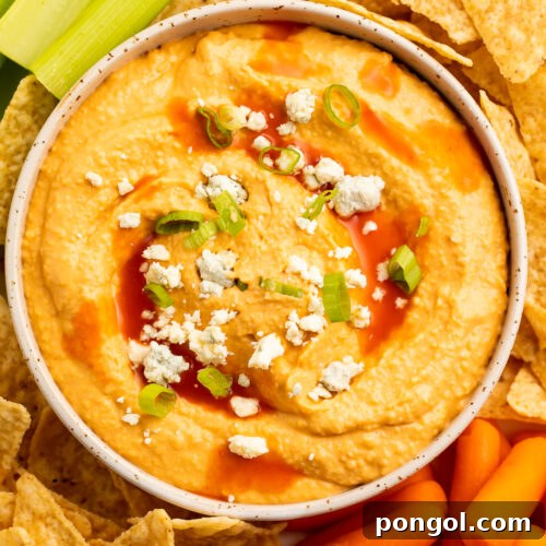 Close-up, overhead view of a bowl of buffalo hummus topped with blue cheese crumbles in the center of a platter surrounded by celery, carrots, and tortilla chips.