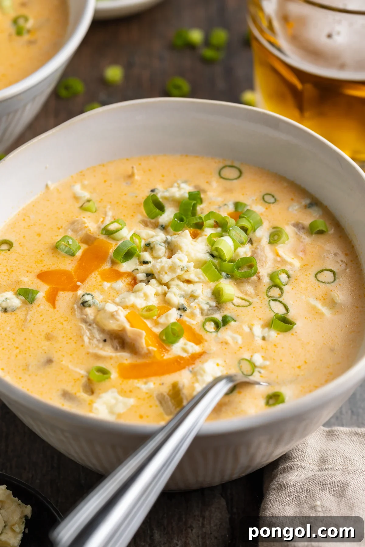 Angled view of a beautiful white bowl filled with pale orange buffalo chicken soup, topped with vibrant green onions and shredded cheese.