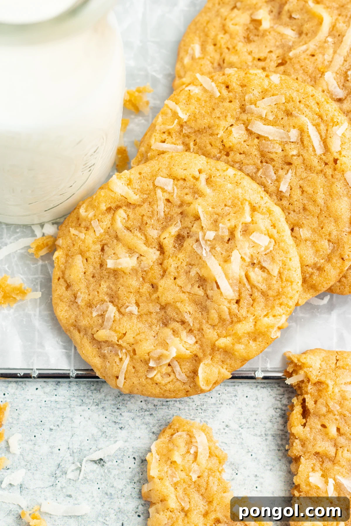 Overhead view of golden-brown coconut cookies on a rustic wooden table next to a refreshing glass of coconut milk, highlighting their inviting texture and color.
