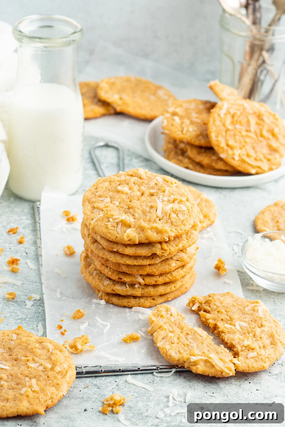 An angled, zoomed-out view of a delicious stack of golden-brown coconut cookies, with a broken cookie showing its soft interior, next to a refreshing glass of milk.
