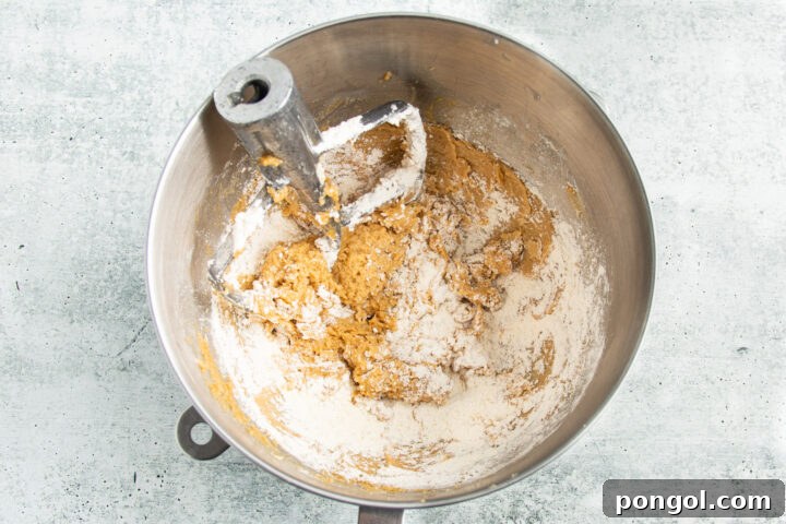 Overhead view of flour mixture being added to the creamed butter and sugars in a large silver mixing bowl, demonstrating the gradual mixing process.