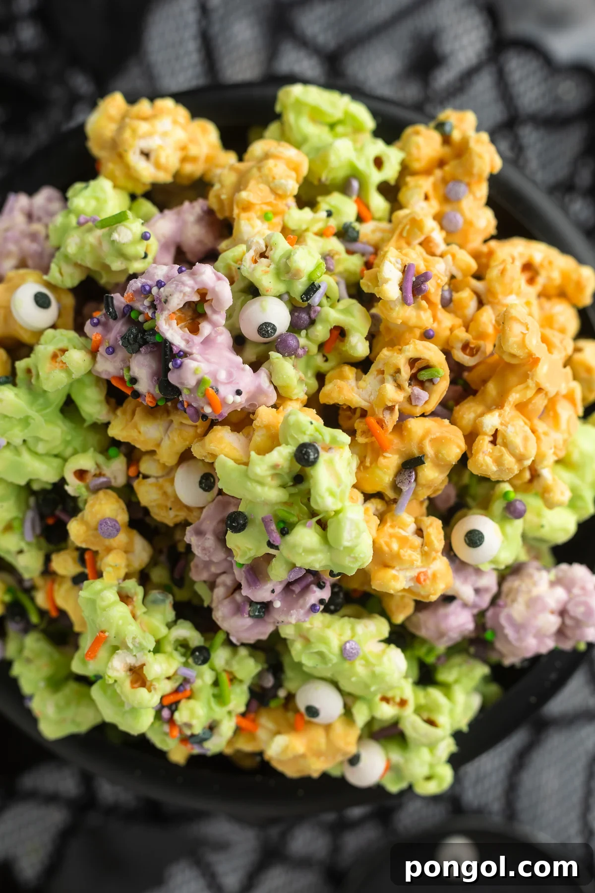 Close-up, overhead view of a plastic cauldron filled with orange, purple, and green white chocolate covered popcorn clusters with sprinkles and candy eyes. This festive popcorn is perfect for Halloween parties or movie nights.
