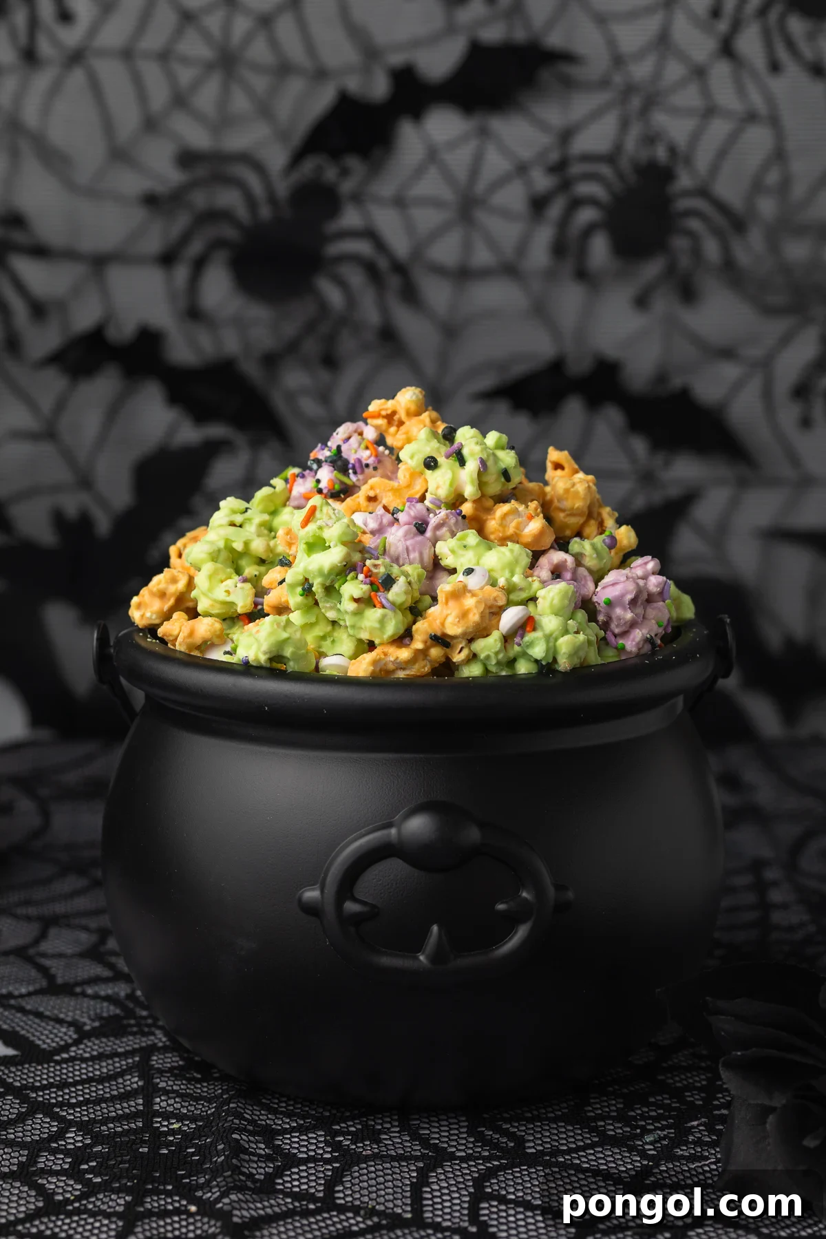 Side view of a plastic cauldron filled with orange, purple, and green white chocolate covered popcorn clusters with sprinkles and candy eyes. This festive popcorn is presented as a Halloween centerpiece.