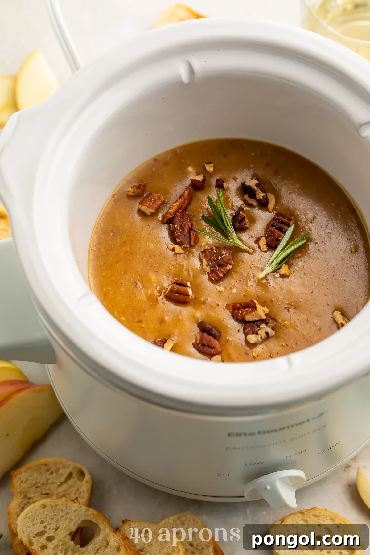 Overhead, angled view of a small white Crockpot slow cooker containing a rich, deep orange baked brie dip topped with crunchy pecans and sweet figs, ready to be served.