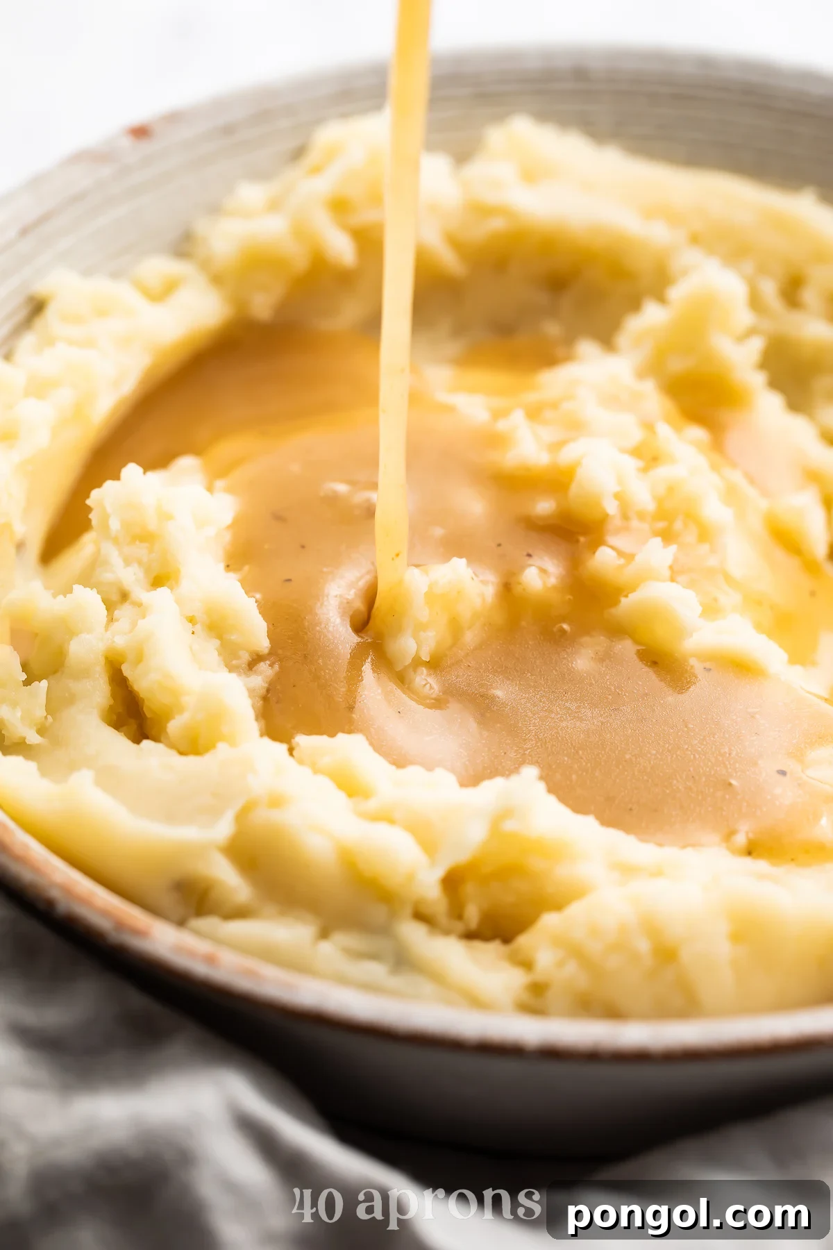 An angled look at a bowl of fluffy mashed potatoes with gluten free gravy being poured on top of the potatoes from above.