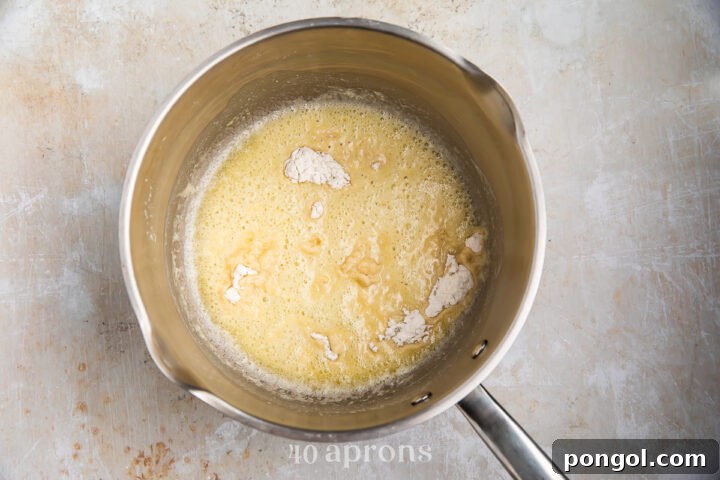 Overhead view of butter and flour for gluten-free gravy in a medium silver saucepan with a handle on a neutral background.