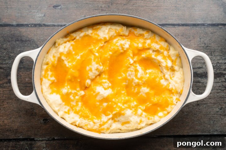 Overhead view of creamy, cheesy mashed potatoes topped with cheddar in a large oval baking dish with handles on each end.
