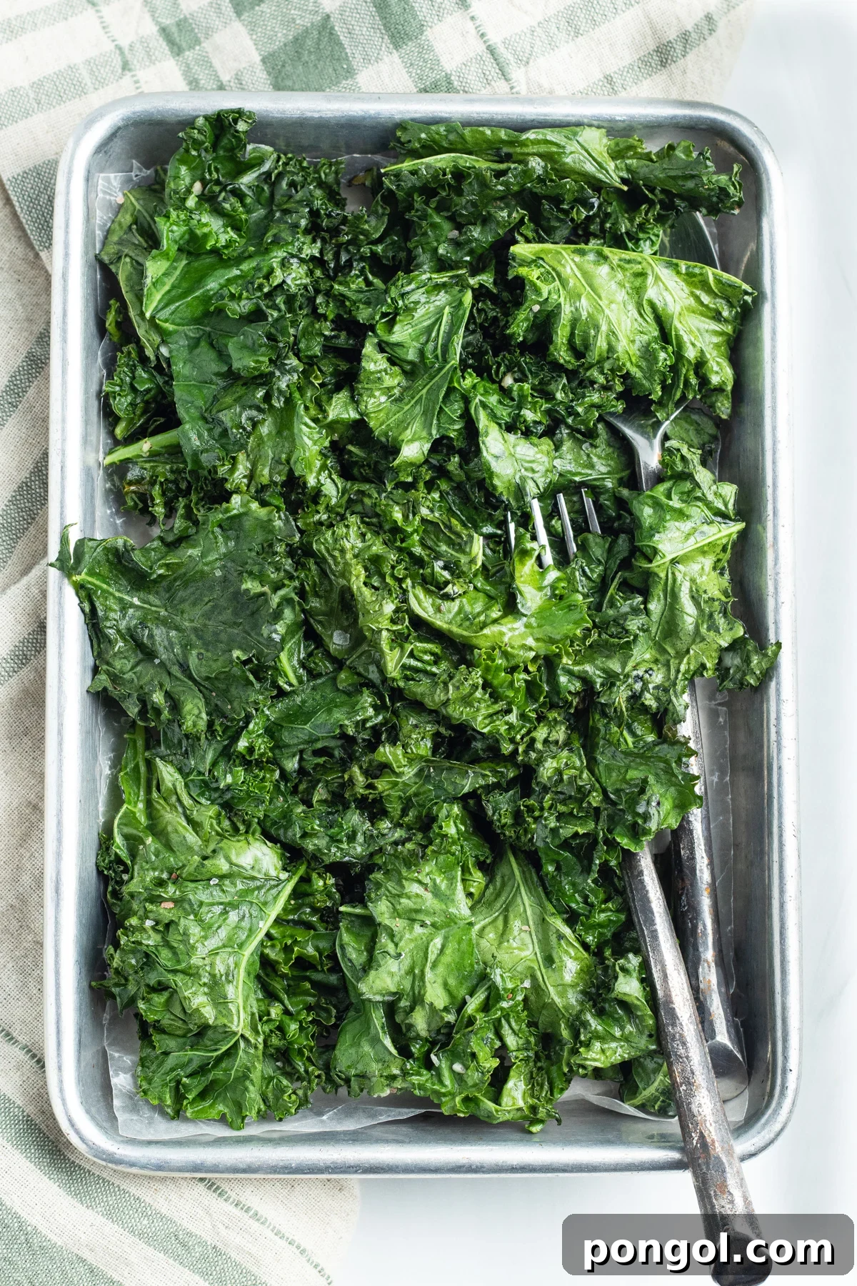 Overhead view of a large sheet pan holding leaves of bright green roasted kale on a neutral tablecloth.