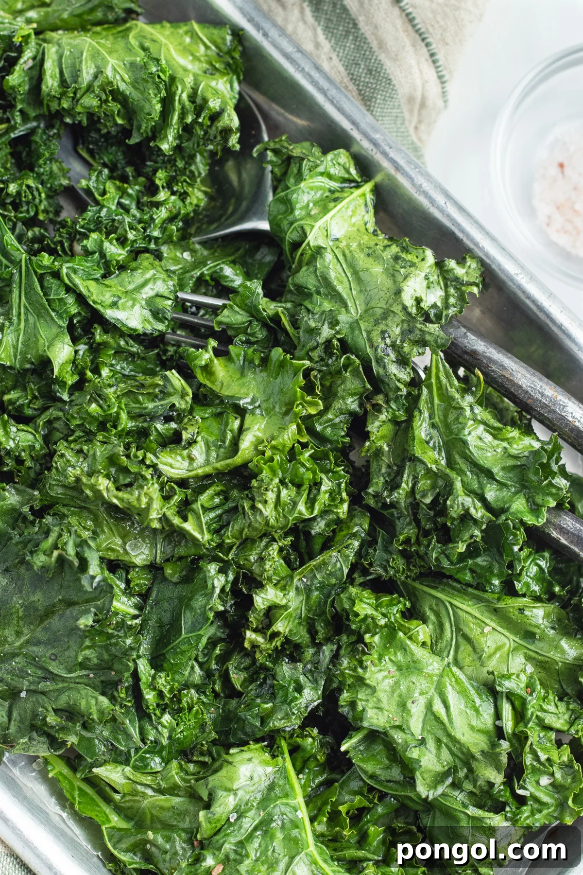 A sheet pan, angled across the field of view, holding bright green leaves of roasted kale with a pair of silver tongs.