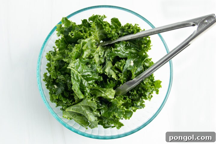 Overhead view of a glass mixing bowl holding large pieces of roughly chopped kale leaves seasoned with oil and garlic.