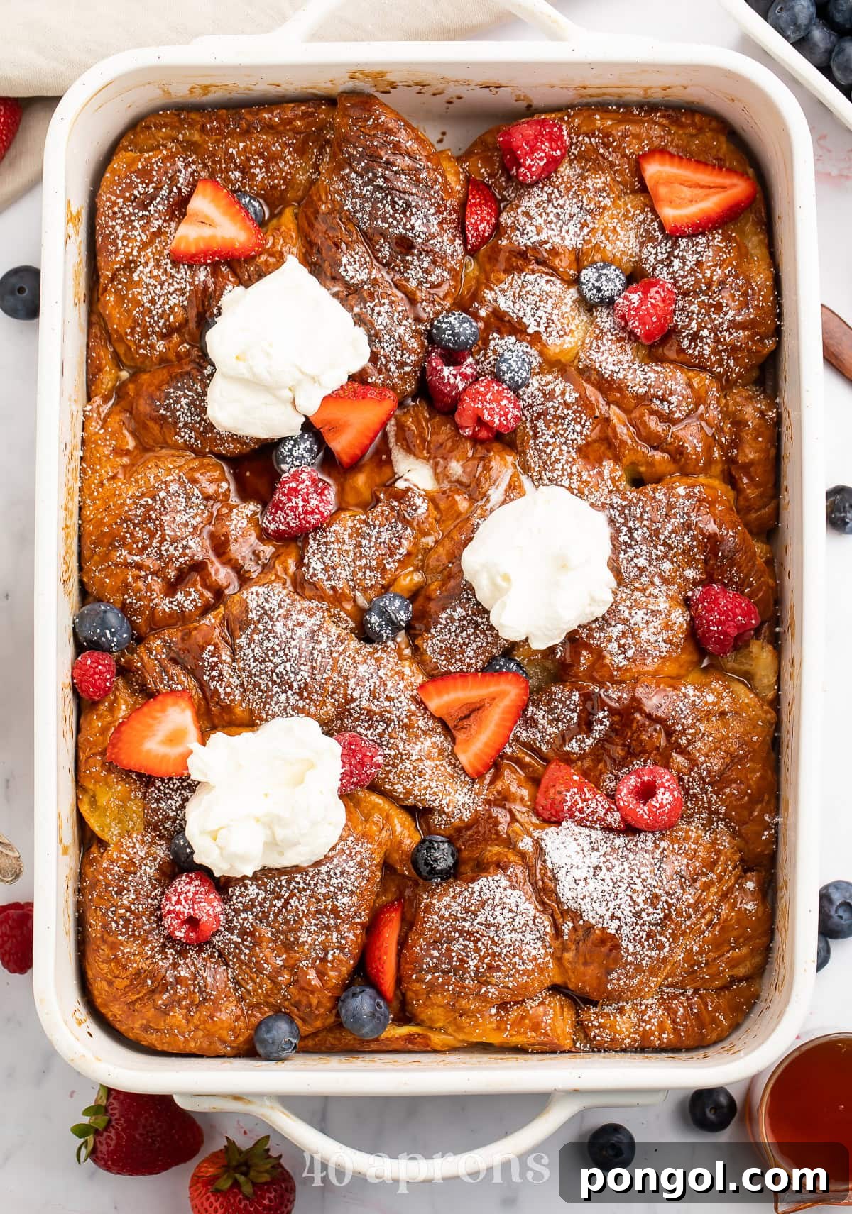 A large casserole dish holding a gorgeous French toast croissant casserole topped with powdered sugar, fresh fruit, and whipped cream.