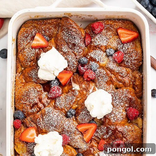 A large casserole dish holding a gorgeous French toast croissant casserole topped with powdered sugar, fresh fruit, and whipped cream.