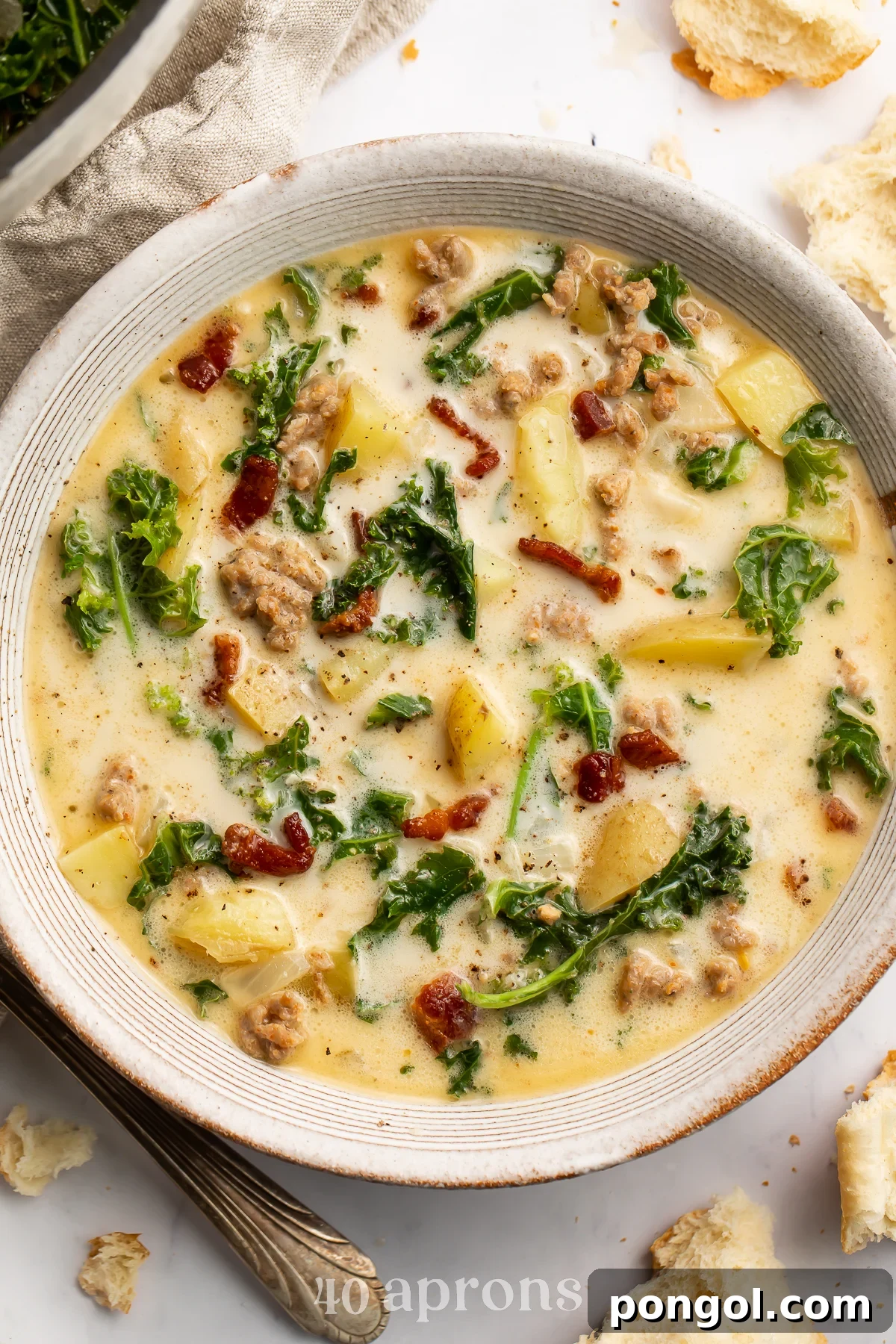 Overhead view of a white soup bowl holding Zuppa Toscana. Floating in the pale yellow soup are spinach, chopped bacon, and chunks of potatoes.