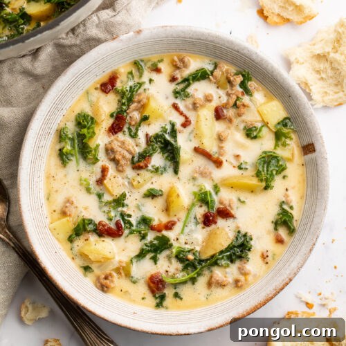 Overhead view of a white soup bowl holding Zuppa Toscana. Floating in the pale yellow soup are spinach, chopped bacon, and chunks of potatoes.