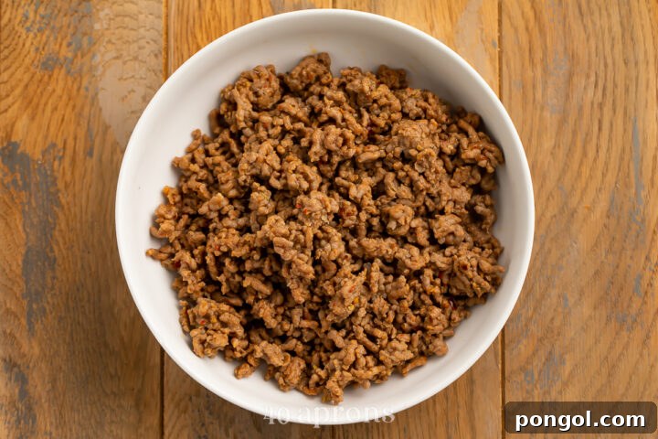 Overhead view of cooked and crumbled Italian sausage in a medium white bowl on a wooden countertop.