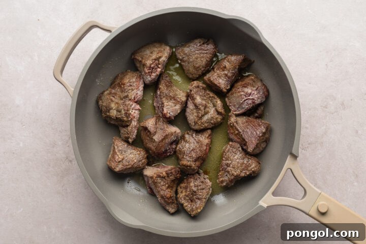 Overhead view of beef tips cooking in a large skillet on a neutral background.