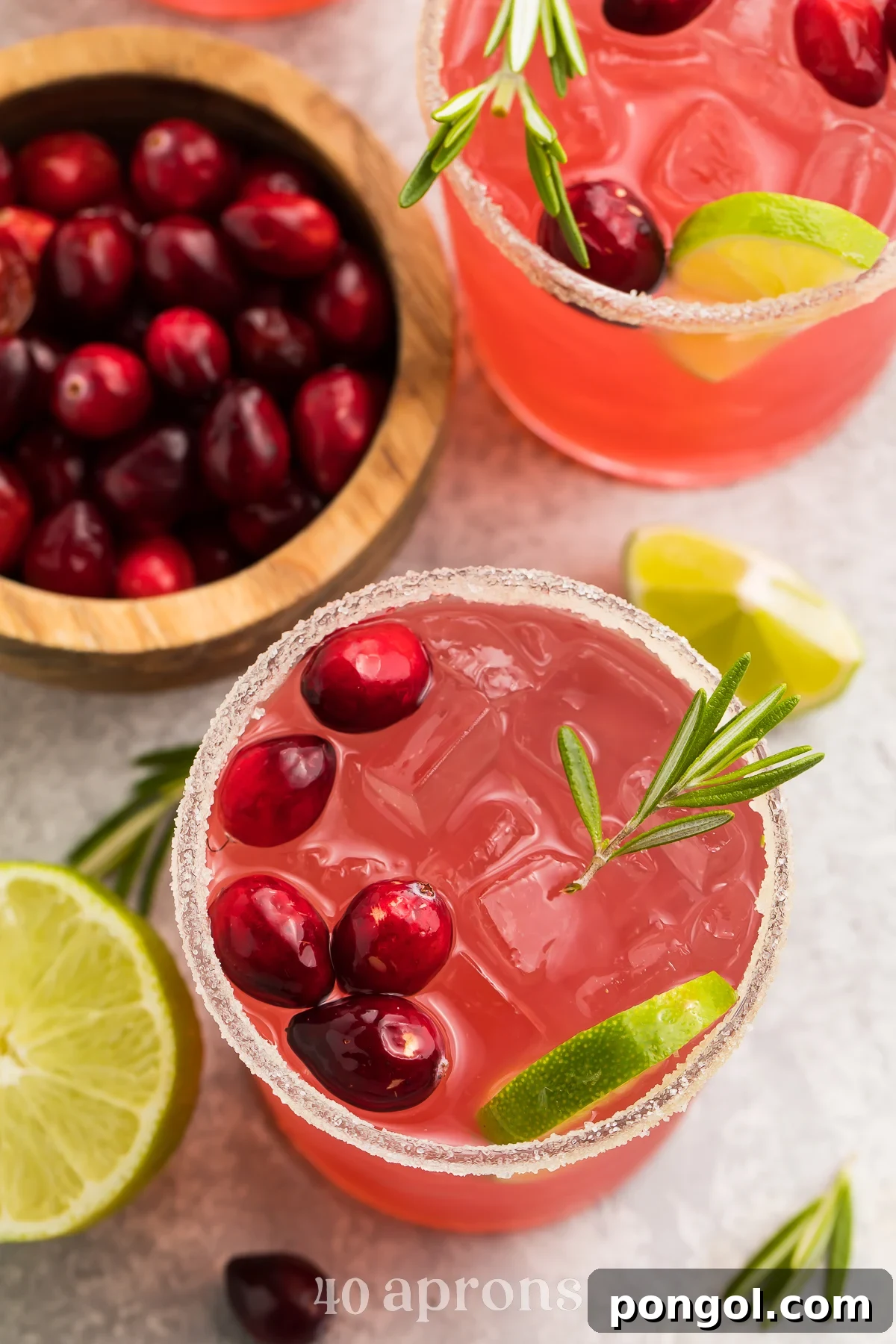 Crimson Margarita 3 Overhead view of several bright red cranberry margaritas arranged on a table. Each drink is elegantly garnished with vibrant green rosemary sprigs and fresh lime slices, highlighting their festive and refreshing appearance.