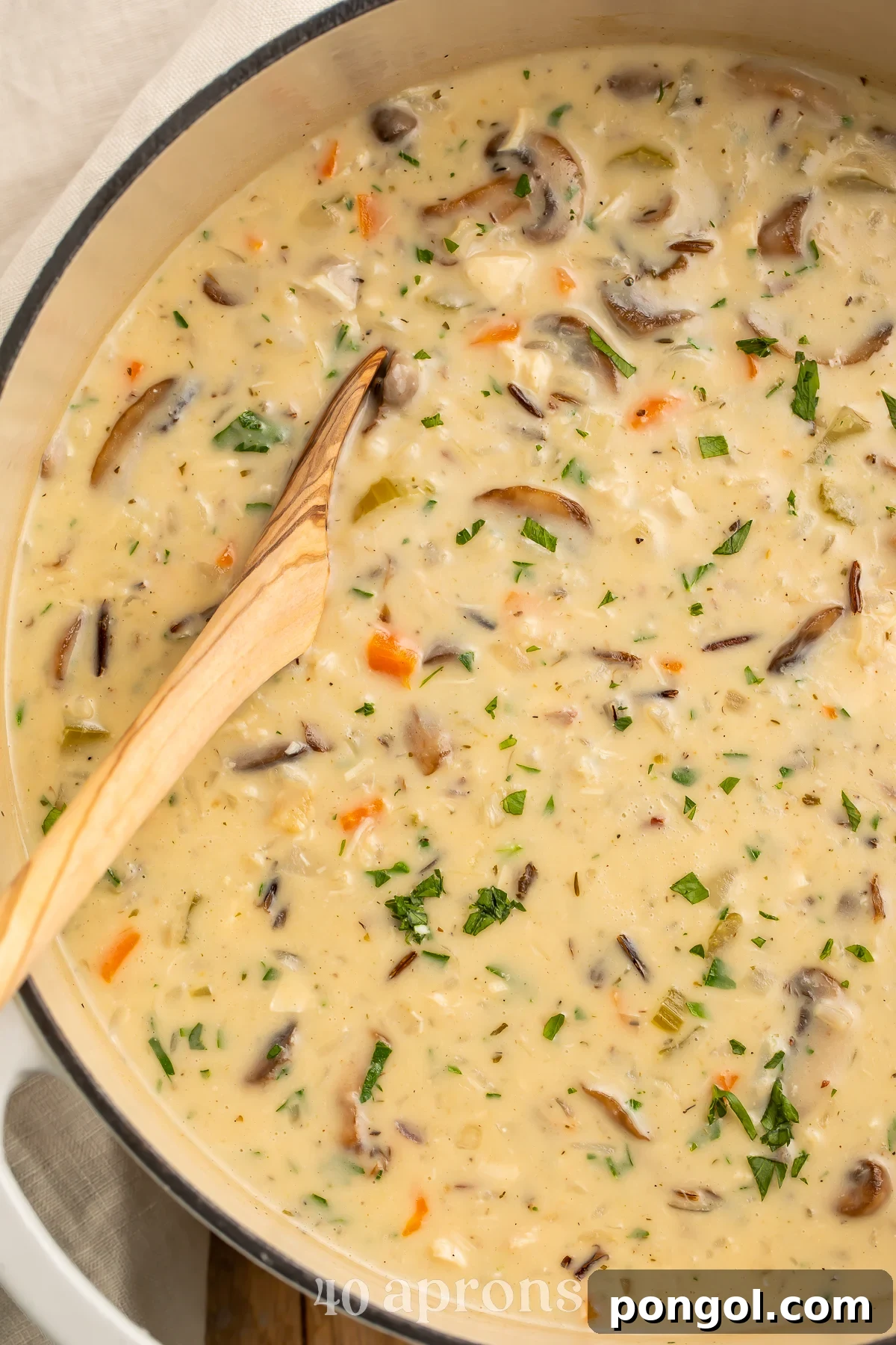 Overhead view of a large heavy pot containing creamy chicken and wild rice soup with mushrooms and carrots. A wooden spoon rests in the soup.