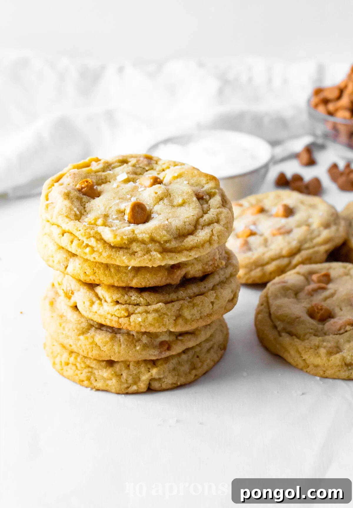 Melt Away Butterscotch Chews 3 A beautifully composed side view of a stack of golden-brown butterscotch cookies, showcasing their thick, soft centers, with more cookies casually arranged in the background next to a white napkin.