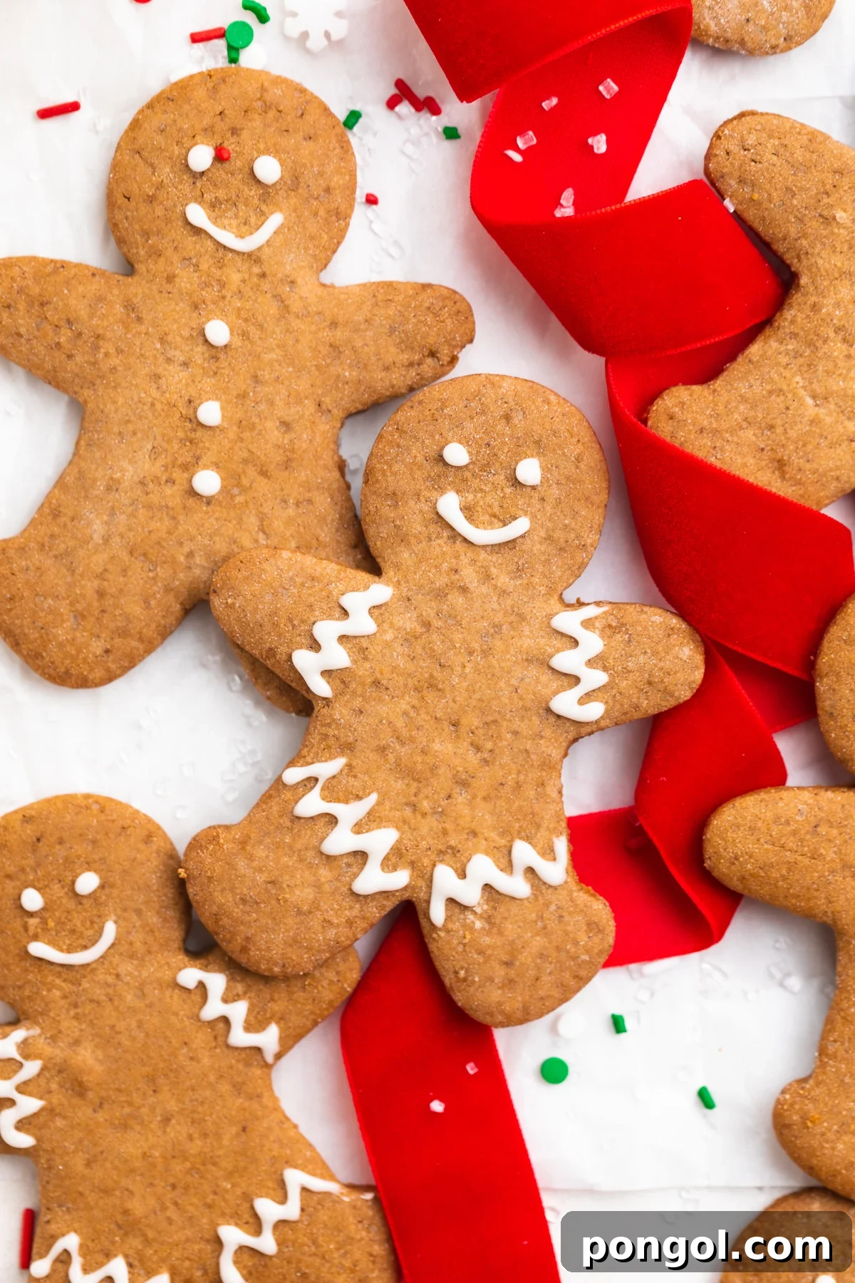 Three beautifully decorated gluten-free gingerbread men cookies, adorned with white icing, resting on a rustic table alongside red holiday ribbon and festive sprinkles.
