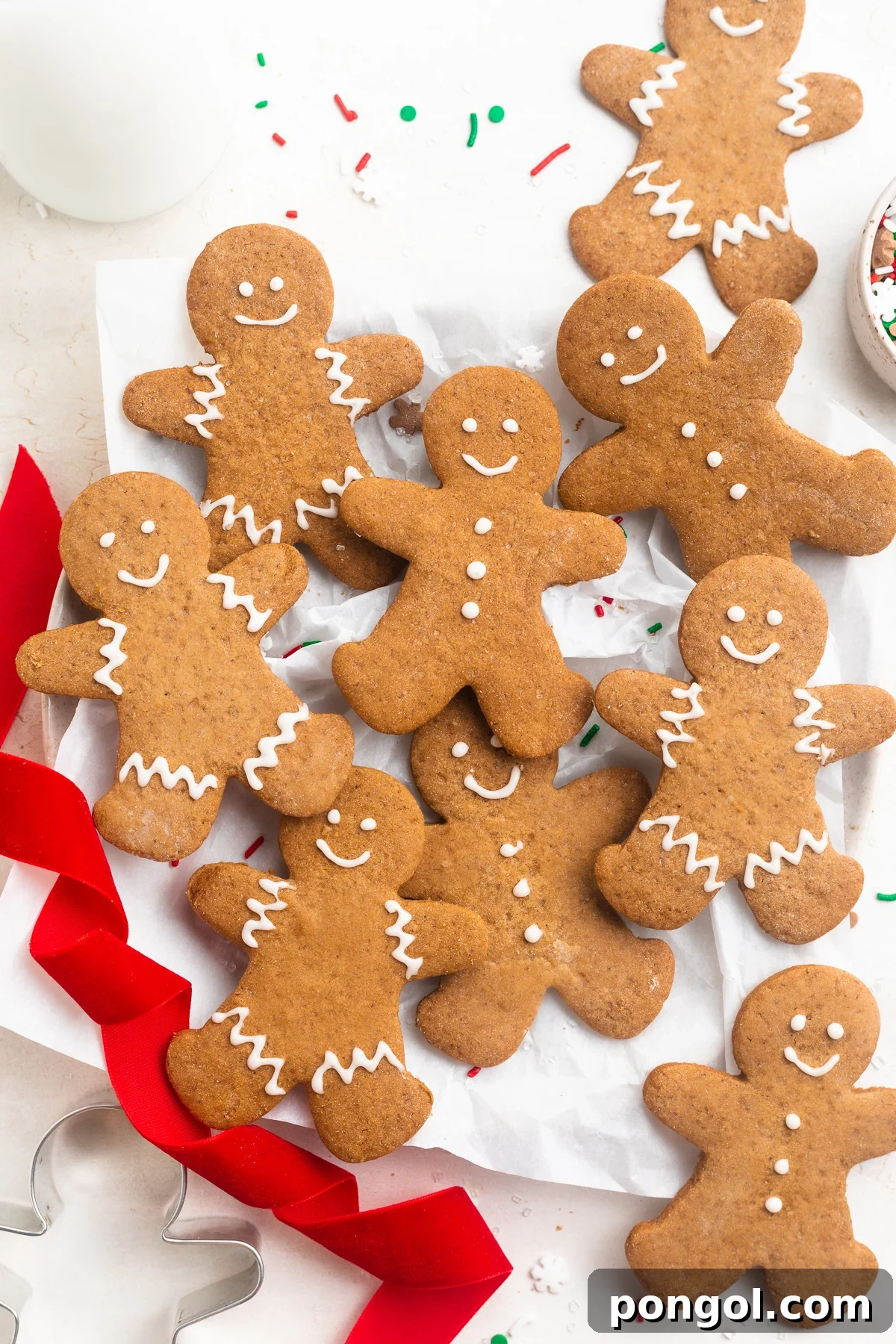 An overhead shot showcasing beautifully decorated gluten-free gingerbread men cookies with white icing, arranged on a white table with festive red ribbon and assorted holiday sprinkles.