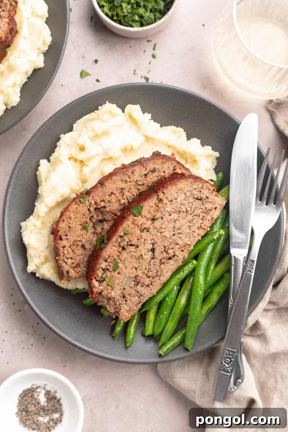 Two slices of Whole30 meatloaf plated with green beans and mashed potatoes on a grey-blue plate with silverware on a table.