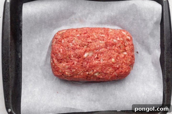Uncooked, unglazed Whole30 meatloaf made with ground beef, shaped into a rectangular loaf in the center of a sheet pan covered in parchment paper.