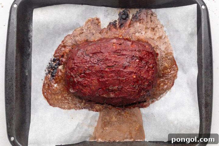 Fully baked, dark brown Whole30 meatloaf on a sheet pan covered with a sheet of parchment paper that's stained from the glaze and beef juice.