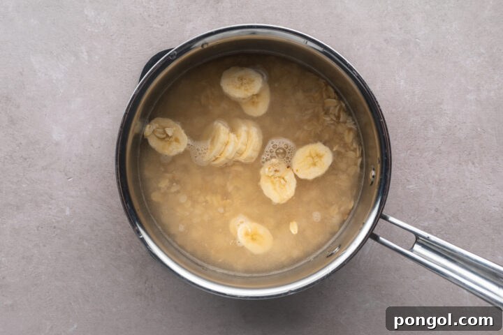 Top-down look at a silver saucepan containing uncooked rolled oats under water, with banana slices floating on top.
