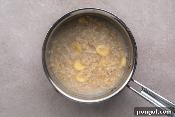 Cooked rolled oats with slices of banana mixed throughout in a silver saucepan with a single handle.