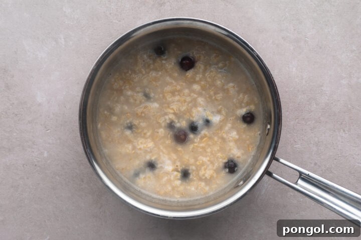 Cooked oatmeal with some water and blueberries in a silver saucepan with a single handle.