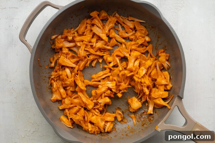 Shredded jackfruit simmering in buffalo sauce in a large skillet, becoming flavorful and tender.