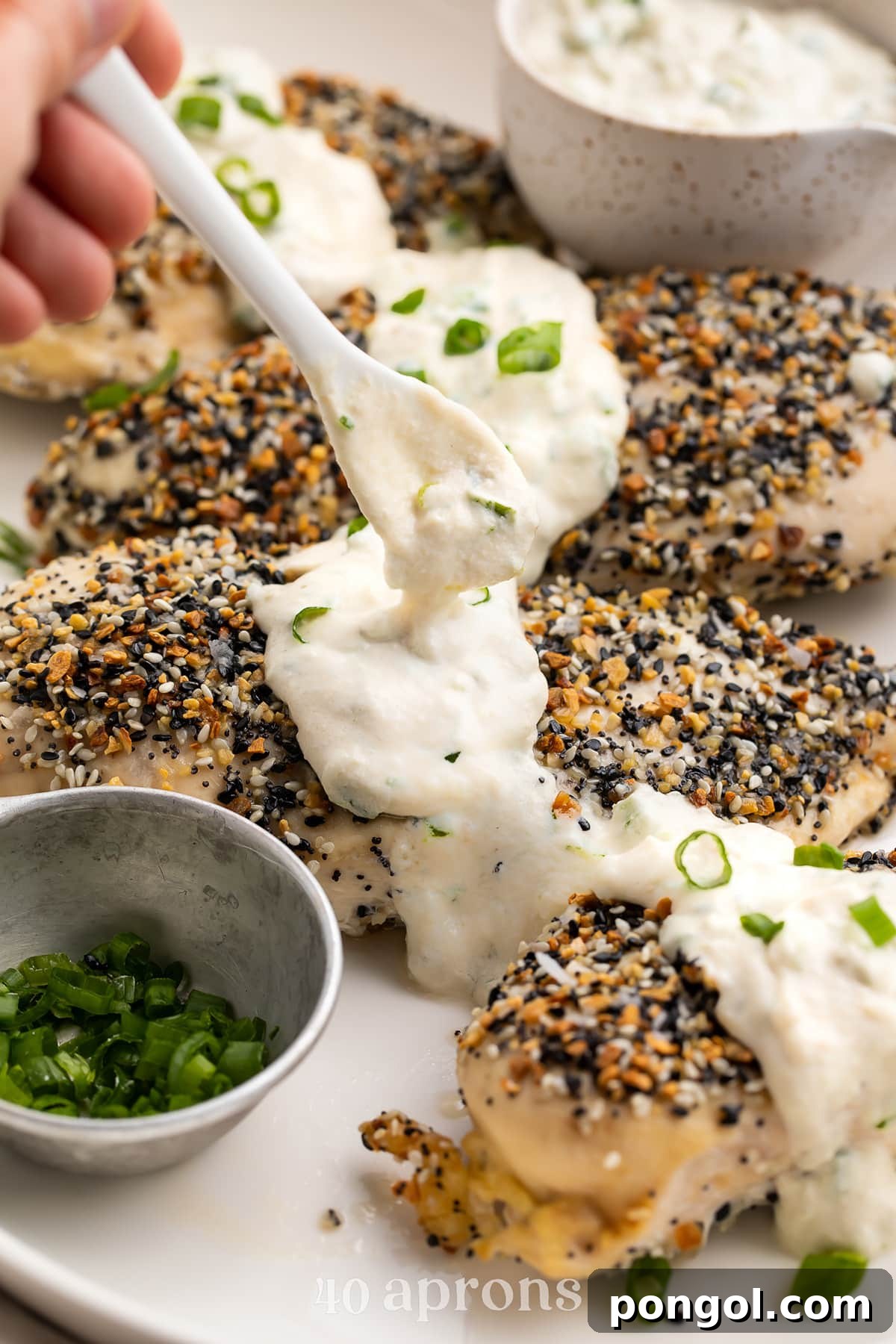 Dairy-free scallion 'cream cheese' sauce being spooned over perfectly baked Whole30 Everything Bagel Chicken breasts in a white dish.