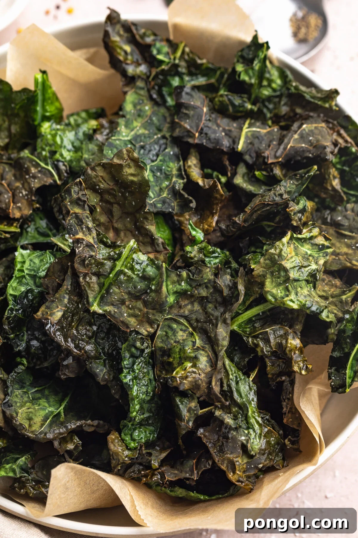 The Ultimate Air Fryer Kale Crunch 2 A large bowl, lined with a sheet of tan parchment paper, filled with crispy crunchy deep green kale chips cooked in the air fryer.