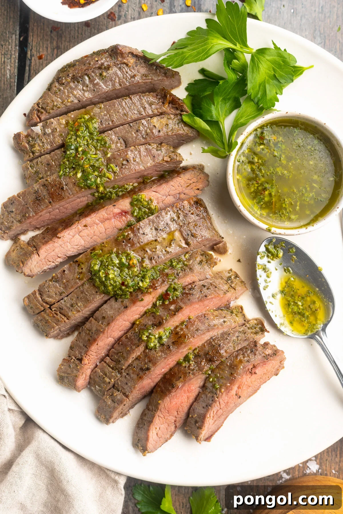 Overhead view of a perfectly cooked sous vide flank steak, thinly sliced and artfully arranged on a white platter, generously garnished with fresh green chimichurri sauce, with an additional ramekin of chimichurri on the side.