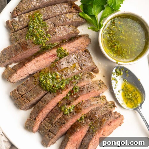 Overhead view of a sous vide flank steak, sliced thin and arranged on a white platter and topped with fresh green chimichurri sauce, next to a ramekin of chimichurri.