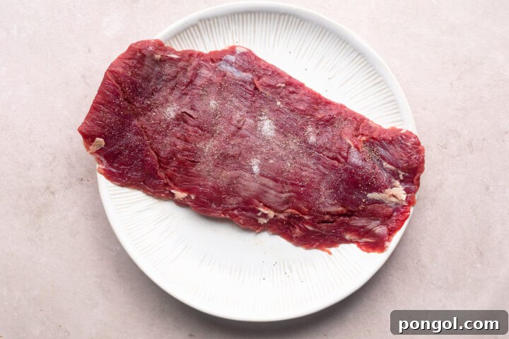 Raw flank steak on a large white plate, seasoned with salt and pepper.
