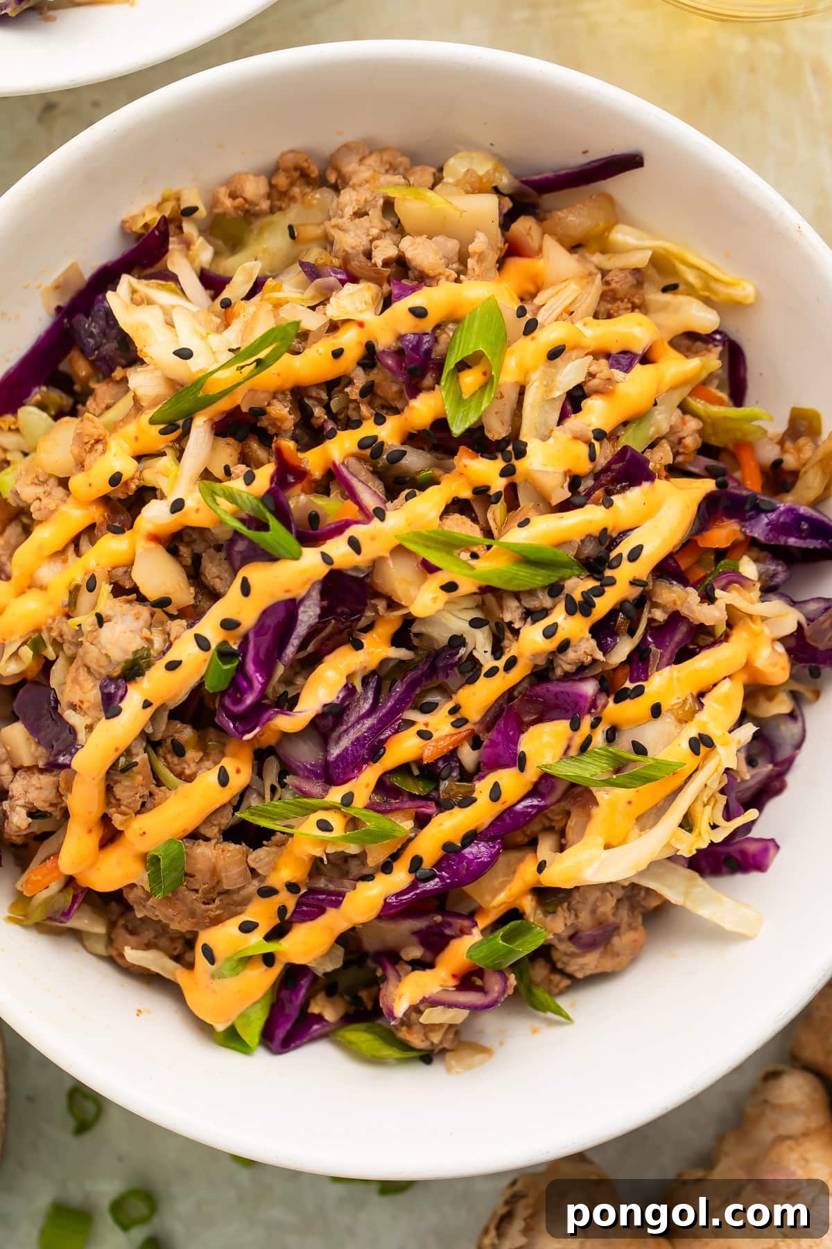 Close-up of egg roll in a bowl with ground pork, purple cabbage, and green onions drizzled with spicy orange sauce and black sesame seeds